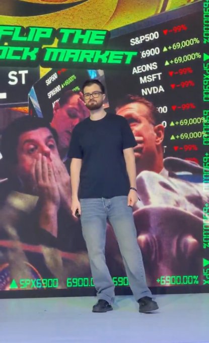 A man with glasses stands on a stage in front of a large screen displaying 'FLIP THE [STOCK] MARKET' and stock tickers like S&P500, MSFT, and NVDA with dramatic percentage changes, alongside images of shocked people.