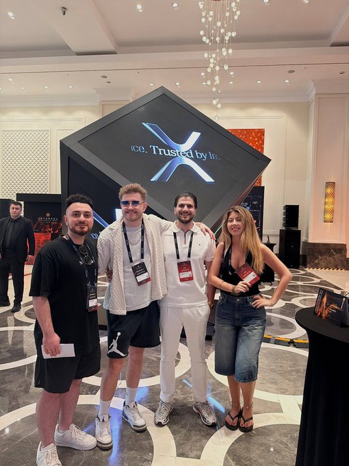 Four people posing for a photo in front of a large screen displaying the X logo and 'Trusted by In...' text at what appears to be a conference.