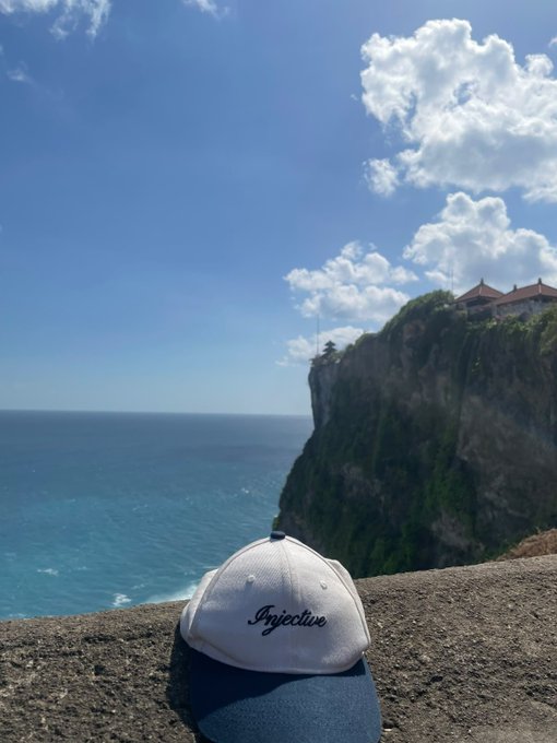 A white baseball cap with 'Injective' written on it sits on a stone ledge overlooking a bright blue ocean and dramatic cliffs topped with a building, under a partly cloudy sky.