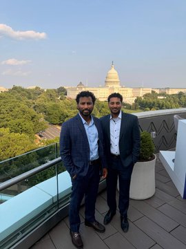 Two men in suits pose on a rooftop terrace with the U.S. Capitol building visible in the background.