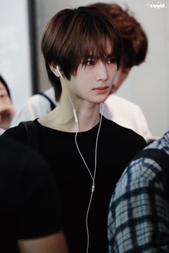 A young man with brown hair wearing earphones and a black shirt looks directly at the camera, likely at an airport arrival as suggested by the ICN airport code in the caption referencing Zhou Anxin's (안신/安信) arrival at Incheon International Airport en route to an Alpha Drive One event.