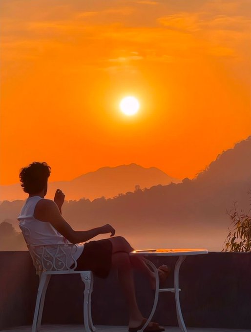 A person sits at a small table on a balcony, watching a bright orange sunset over misty mountains.
