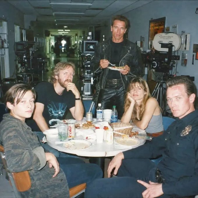 A behind-the-scenes photo from the set of Terminator 2 showing Edward Furlong, James Cameron, Linda Hamilton, Arnold Schwarzenegger, and Robert Patrick eating together around a table loaded with food and a visible Coca-Cola can.