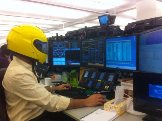 A man wearing a yellow motorcycle helmet sits at a trading desk on the GS (Goldman Sachs) trading floor, surrounded by multiple monitors displaying financial data and charts as he types on a keyboard. The image captures his playful preparation for a Harlem Shake video, capturing the lighthearted moment on the trading floor before compliance intervened.