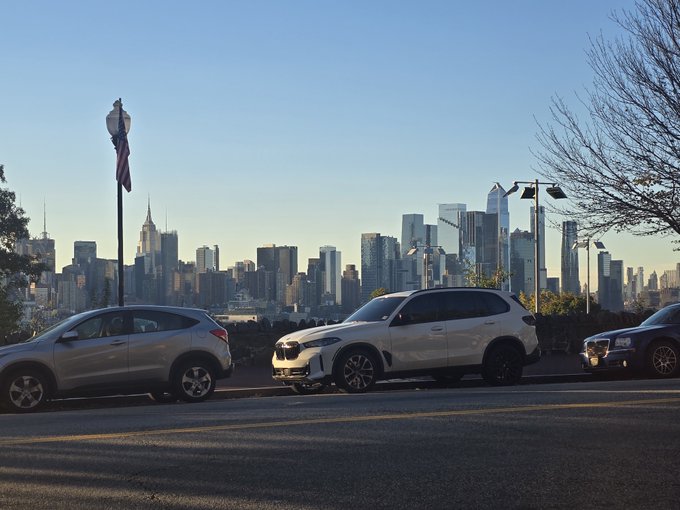 Three vehicles are parked in the foreground overlooking the Manhattan skyline, which is silhouetted against a clear blue sky during the daytime.