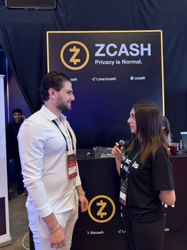 In this image, a man wearing a white shirt stands next to a woman who is holding a microphone. They are both facing each other at an event where they appear to be talking or presenting something related to cryptocurrency. The background of the image features a black curtain and a sign that says "Zcash". There are also some people in the background, possibly attending the same event as well.
