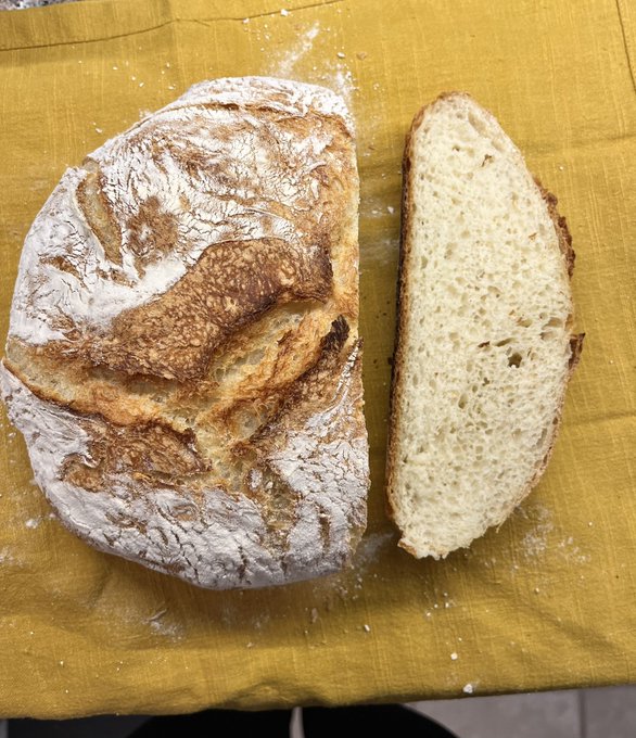 A freshly baked loaf of crusty sourdough bread sits on a golden-yellow cloth, with one thick slice cut and laid beside it, revealing a soft, airy crumb—perfect inspiration for "getting this bread" to start the day.