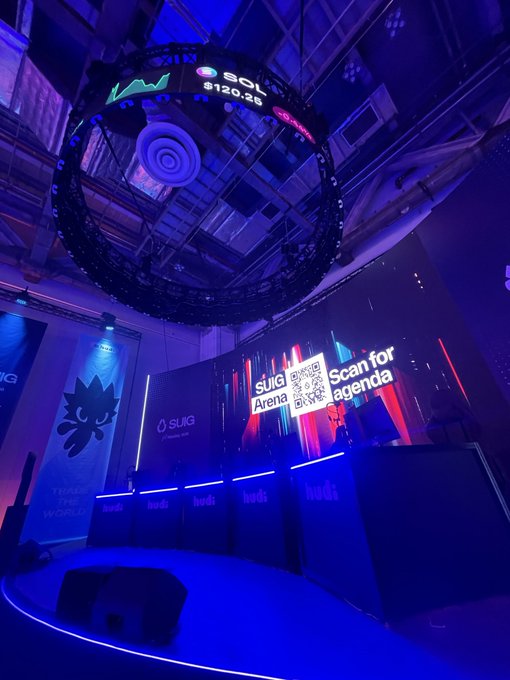 A futuristic event space at Token2049 Basecamp MBS glows in deep blue lighting, featuring a large circular overhead display showing "$SOL $120.25" and a prominent screen prompting viewers to "Scan for agenda" with a QR code labeled "SLUG Arena." Branding for "SLUG" and "hudd" lines the booth, tying into the post's announcement of a Huddy social trading competition starting at 11:30 AM.