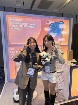 Two young women pose at a convention booth with an orange backdrop titled 'Markets Without Borders: Trade, Global Financial Markets,' one holding a small panda plush toy.