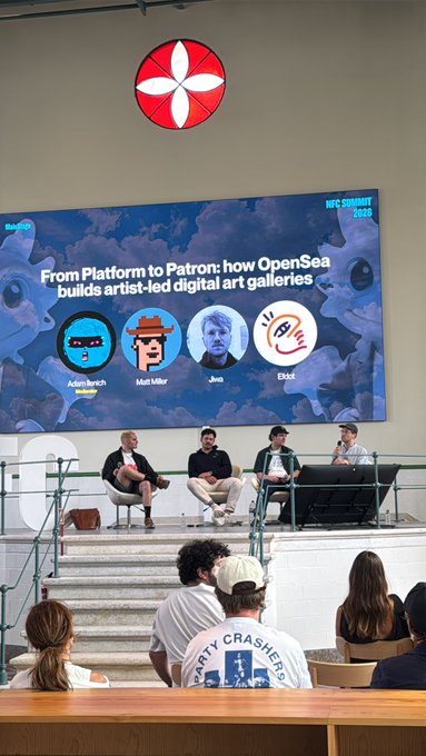 The image shows a group of people sitting around a table, with some standing behind them. They are gathered for an event or discussion, possibly related to the OpenSea Gallery. The room has a large blue wall featuring a red circle with four white faces inside, which is likely part of the event setup. A banner can be seen hanging on the wall as well.