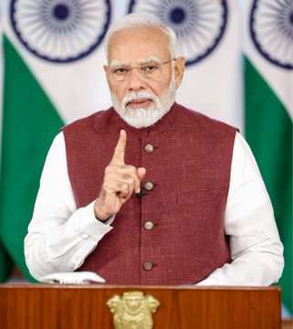 Indian Prime Minister Narendra Modi speaking at a podium with the Indian flag and Ashoka Chakra visible in the background, raising his index finger.