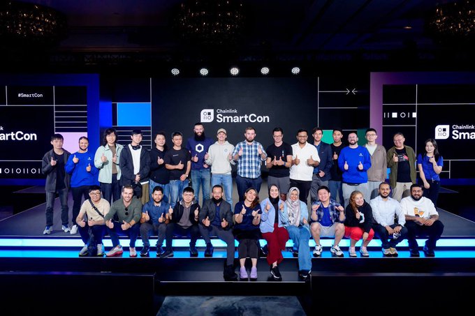 A large group of people pose together on a tiered stage at "Chainlink SmartCon," with the event branding visible on screens behind them.