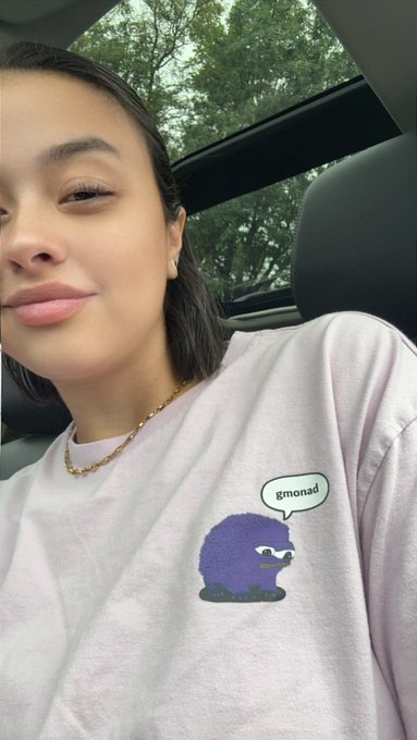 A young woman with short dark hair is sitting in a car wearing a white t-shirt with a purple cartoon character and a speech bubble saying 'gmonad'.