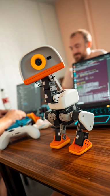 A small robotic figure with orange and white components stands on a wooden desk in front of a blurred man working on a laptop displaying code.