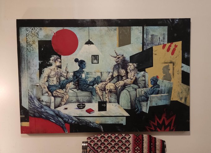 A painting hanging on a beige wall depicts surreal figures sitting on couches in a living room, with a red sun, a horned creature, and various abstract elements; a patterned blanket is visible below the artwork.