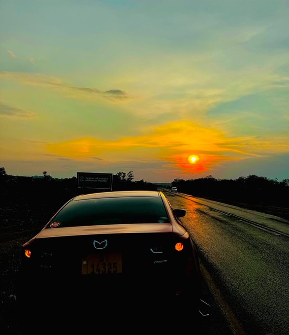 A scenic photograph of a white Mazda RX-7 parked on the side of a highway during a vibrant sunset, with the car's brake lights illuminated.