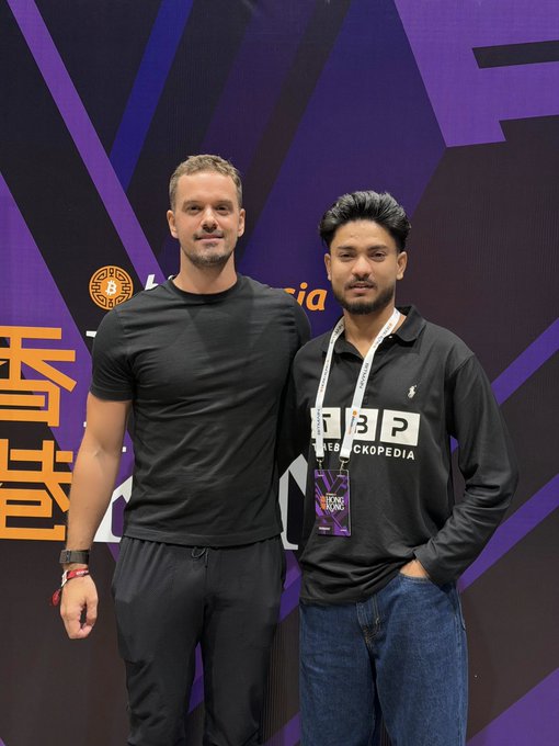 Two men pose together at what appears to be an event, one in a fitted black t-shirt and the other wearing a black "DROP" shirt with a lanyard.