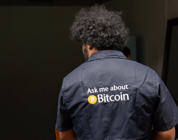 A man with curly hair seen from behind wearing a dark blue shirt that reads 'Ask me about Bitcoin' with an orange Bitcoin logo.