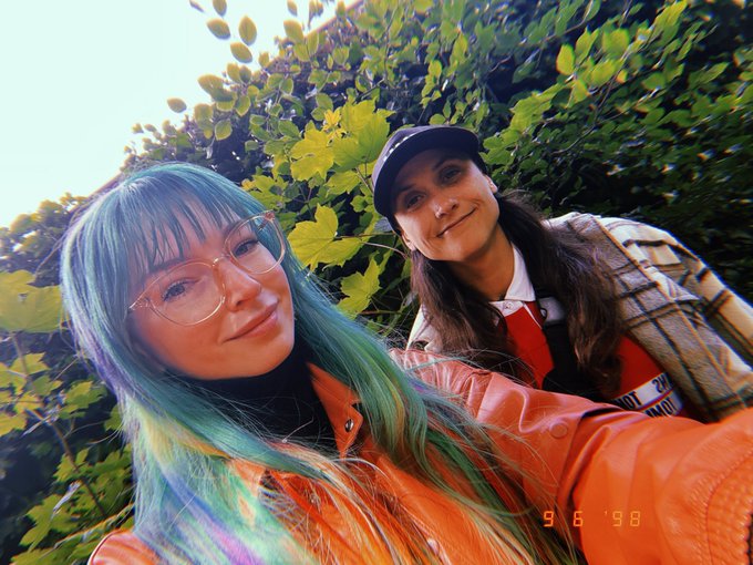 A retro-styled photo dated 9/6/98 of two people outdoors against green foliage, one with long blue and green hair wearing glasses and an orange jacket, the other wearing a cap and plaid shirt.