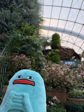 A light blue, sad-faced cartoonish figurine stands in the foreground of a greenhouse with a glass ceiling.