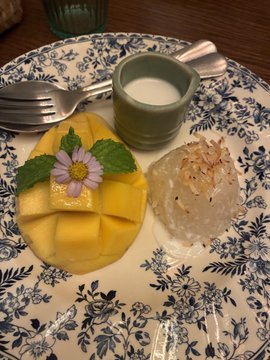 A decorative blue and white patterned plate containing sliced mango topped with a purple flower, alongside a small cup of what appears to be coconut milk and a dessert topped with shredded coconut.