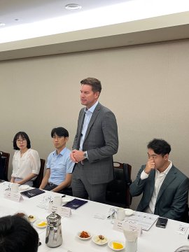 A man in a gray suit stands addressing a seated group during a closed-door meeting in Seoul, with a woman and two other men listening attentively around a conference table set with refreshments. The gathering reflects the post's description of Optimism's sit-down with the Ethereum Foundation and Korea's financial and policy leaders, organized by @ethereumkoreaio ahead of September's Ethereum flagship conference.