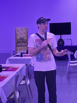 A man wearing a baseball cap and a graphic t-shirt holding a microphone, appearing to speak or host an event in a studio with purple lighting, with merchandise visible on tables nearby.