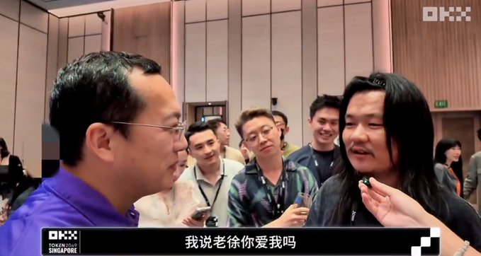 A group of people gather around a man with glasses at the TOKEN2049 Singapore event, with one person asking "我说老徐你爱我吗" (I said, Lao Xu, do you love me?) as shown in the OKX-branded subtitles, capturing an interview moment during the crypto conference.