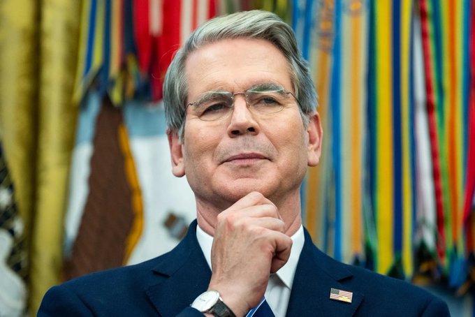 A portrait of a man wearing glasses and a suit with an American flag pin, standing thoughtfully with his hand on his chin in front of various flags.