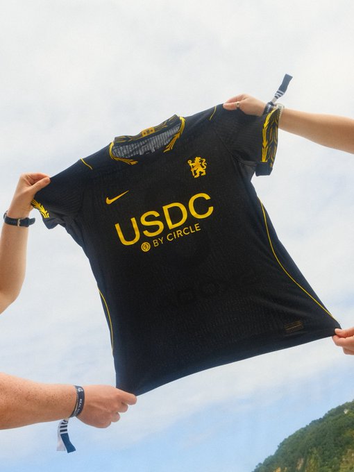 Two people holding up a black and yellow sports jersey featuring the text 'USDC BY CIRCLE' and a Nike swoosh against a cloudy sky background.