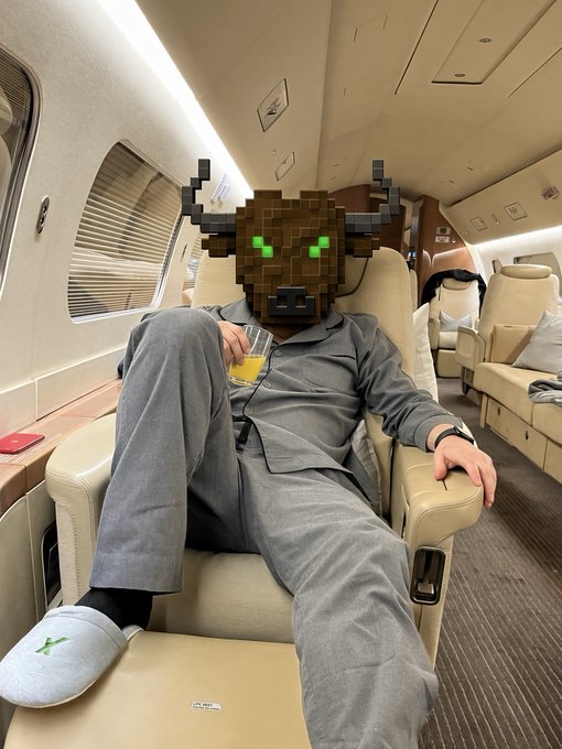 A person wearing gray pajamas and slippers sits in a luxurious airplane seat holding a glass of orange juice, with a Minecraft-style pixelated creature filter applied to their face.