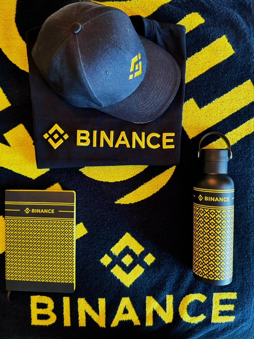 A flat lay display of Binance merchandise including a branded cap, water bottle, notebook, and blanket with the yellow Binance logo.