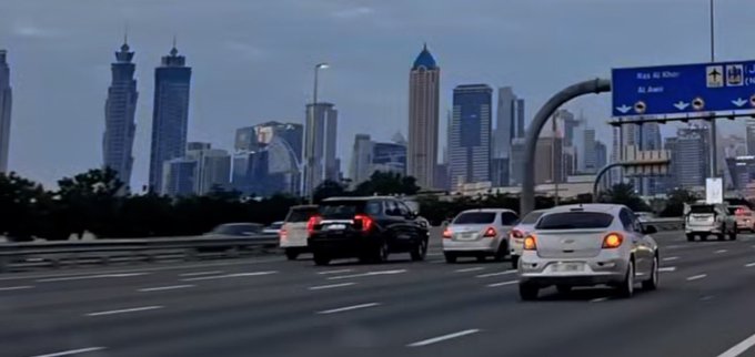 iced glass skyscrapers rise against a hazy blue sky, with cars and a street sign visible on the right side of the frame.