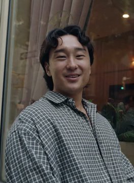 A casual portrait photograph of a smiling young East Asian man with dark wavy hair, wearing a checkered button-up shirt, posed indoors near a glass wall with curtains.
