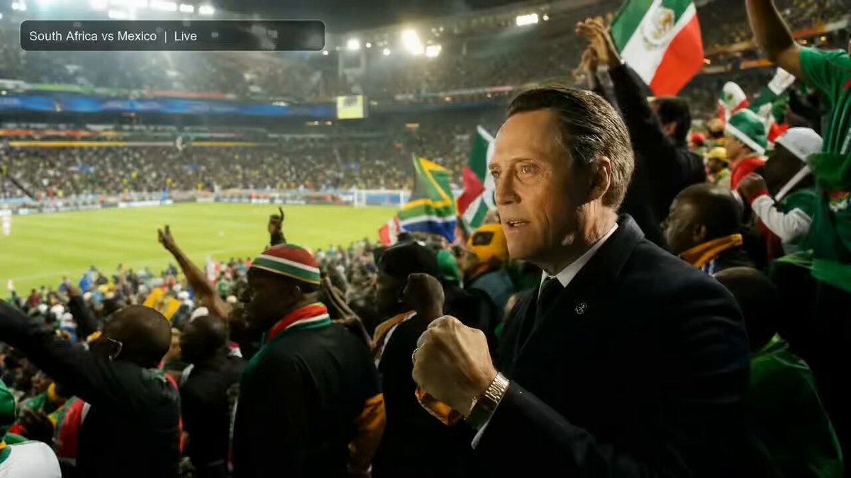 The image shows a man standing at a soccer stadium, surrounded by a large crowd of people who are cheering for their team. The man appears to be wearing a suit and tie, which contrasts with the casual attire of the fans around him. The scene captures the excitement and energy of a live soccer match as the spectators show their support for their favorite teams.