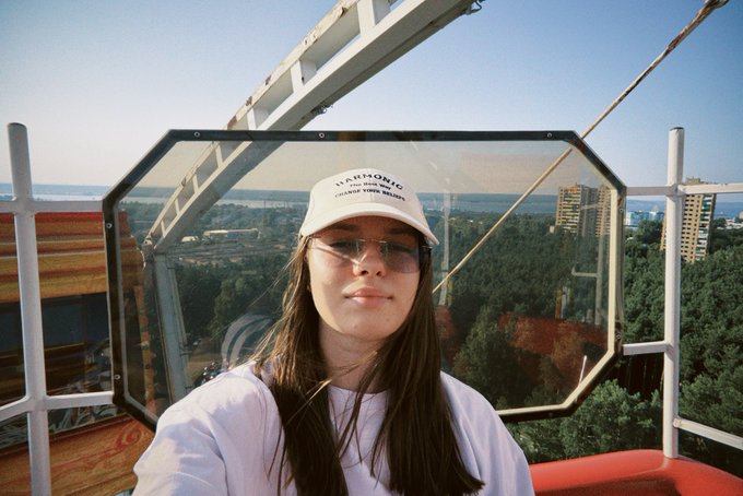 A selfie of a young woman wearing sunglasses and a white cap, taken from what appears to be a Ferris wheel overlooking a city and water.