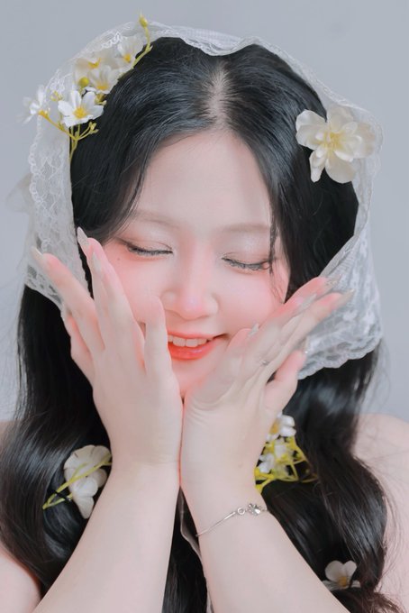 A portrait photo shows a young woman with long dark hair wearing a lace veil adorned with white flowers, smiling with her eyes closed and hands near her face, wearing long manicured nails and a delicate bracelet.