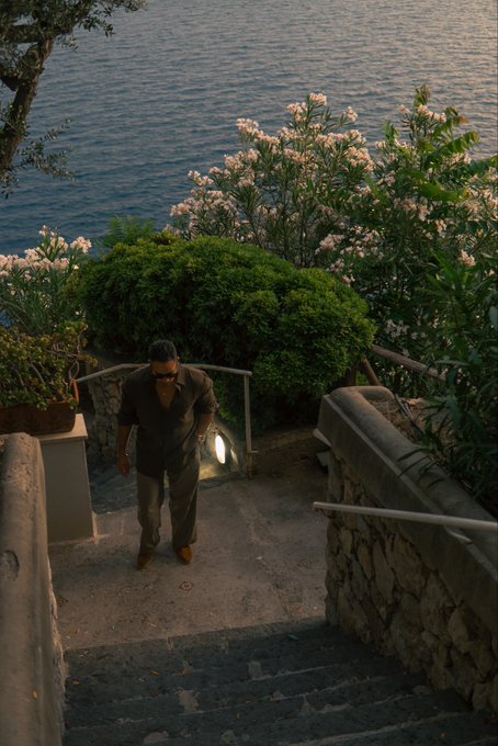 A man in a dark shirt and sunglasses walks up stone stairs from a coastal garden adorned with blooming white flowers. The serene seaside setting complements a peaceful "Good morning fam" greeting.