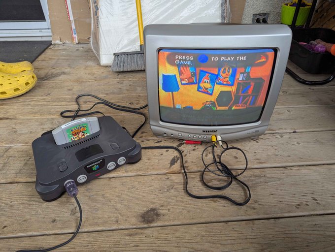 A Nintendo 64 console is connected to a small CRT television displaying the title screen for "Banjo-Kazooie," which reads "PRESS [button icon] TO PLAY THE GAME." The setup sits on a wooden floor with a broom and other items visible in the background.