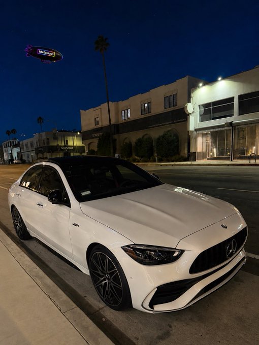 The image features a white Mercedes-Benz car parked on a city street at night, with a building visible behind it. The car appears to be in good condition and well-maintained. In the background of the photo, there are two buildings - one is a tall structure that stands out against the dark sky, while the other is relatively shorter. A purple balloon can also be seen floating above the cityscape. The image includes text that reads "2028 nights Prob will be a lambo tho $XPR #crypto".