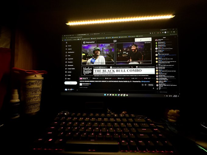 A laptop in a dimly lit room displays a Polymarket livestream featuring hosts promoting "THE BLACK BULL COMBO" from Market Bubble, accompanied by a backlit keyboard and a partially visible coffee cup, illustrating the late-night viewing experience across different time zones mentioned in the post.