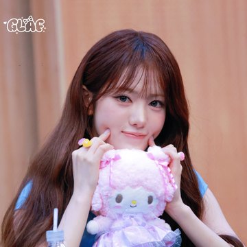 A young woman with long brown hair smiles while holding up a pink Sanrio plush toy (My Melody) with both hands.