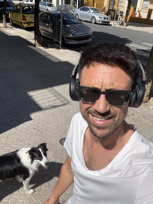 A selfie of a smiling man wearing sunglasses, white t-shirt, and black over-ear headphones, with a black and white dog standing on a paved street behind him.