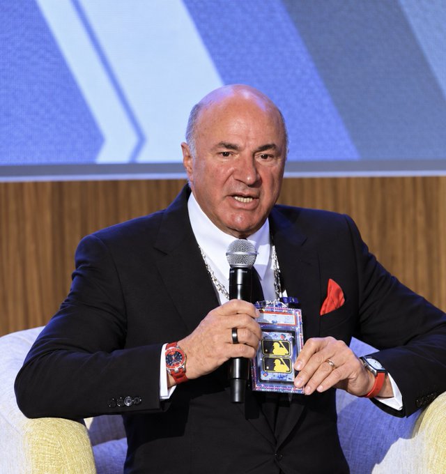A bald man in a dark suit with a red pocket square speaks into a microphone while holding a clear display case containing trading cards.