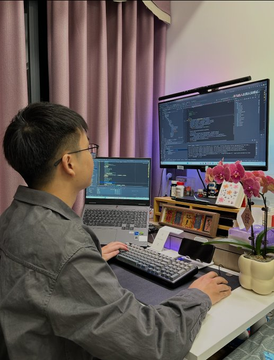 A young man wearing glasses and a gray jacket sits at a desk with a laptop and an external monitor displaying code, in a cozy room with pink curtains and a small vase of flowers.