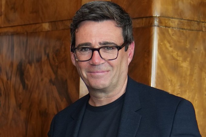 A portrait of a middle-aged man with short dark hair and rectangular glasses, wearing a navy blazer over a dark t-shirt, posed against a wood-paneled wall. The image appears to accompany a post announcing Andy Burnham as the UK's new Prime Minister following Keir Starmer.