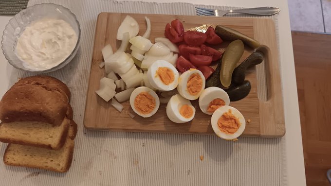 A wooden cutting board with sliced boiled eggs, tomatoes, onions, and pickles, alongside a bowl of white sauce and sliced brown bread.