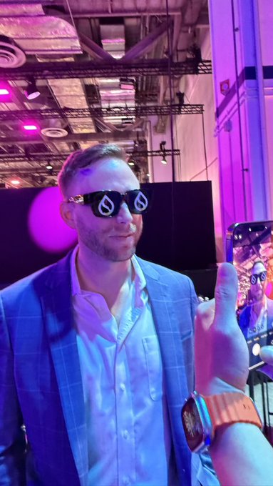 A man in a blue suit wearing distinctive sunglasses with a spiral design poses for a photo while someone takes his picture on a smartphone at an indoor venue with purple lighting.