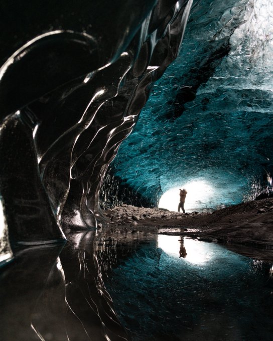A stunning photograph of the inside of an ice cave in Iceland, with a person standing at the entrance silhouetted against the light, and their reflection visible in the water on the ground.