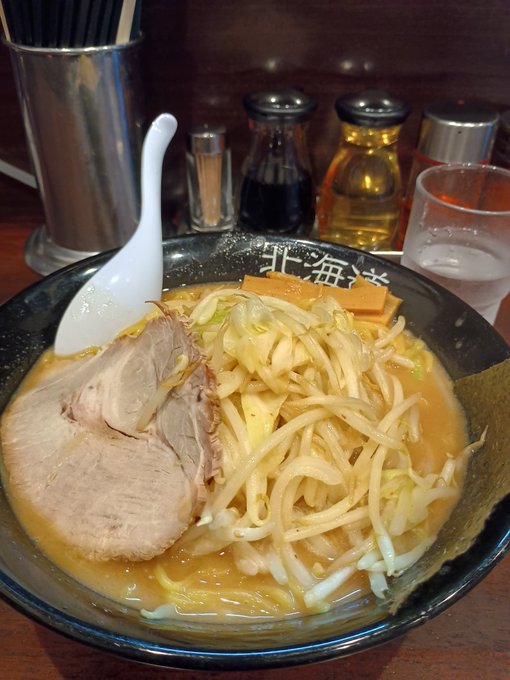 A bowl of ramen with chashu pork, bean sprouts, and other toppings in a dark broth, served at a table with condiments and condiments in the background.